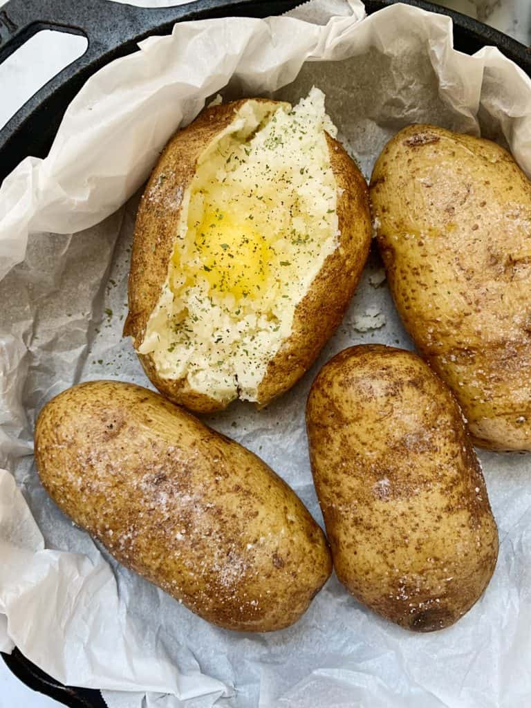 Four Instant Pot Baked Potatoes in a skillet. One is split open with ghee, salt, and pepper.