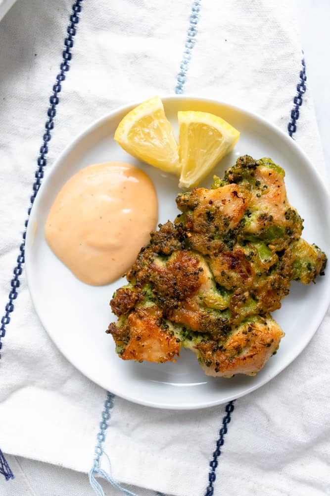 one of the chicken broccoli fritters on a small plate with dipping sauce and lemon wedges