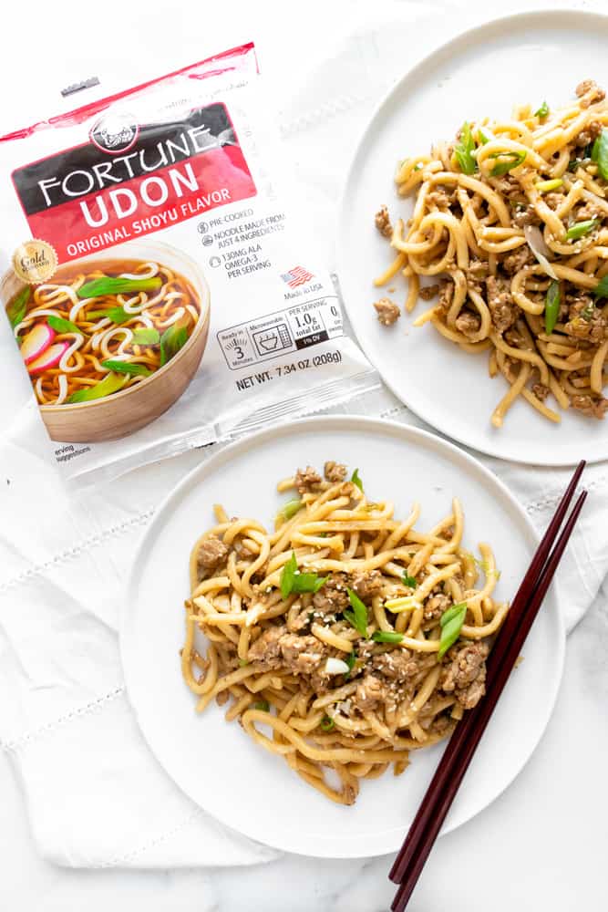 two plates of sesame udon stir fry with the packaging of the udon noodles next to them