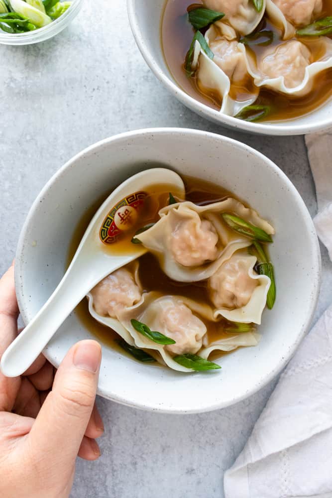 close up of easy wonton soup with a spoon in the bowl and a hand holding the bowl