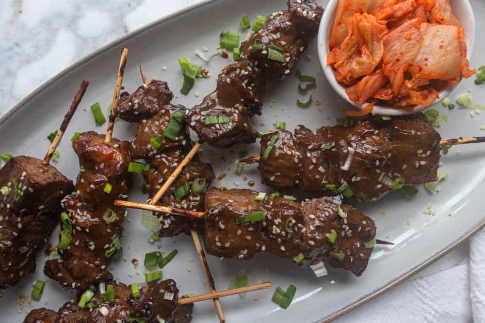 Korean BBQ Skewers on a platter next to a bowl of kimchi